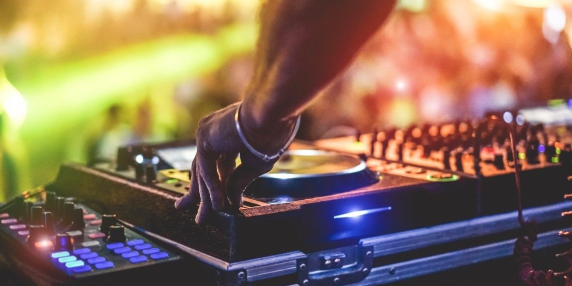 Dj mixing outdoor at beach party festival with crowd of people in background - Summer nightlife view of disco club outside - Soft focus on hand fingers - Fun ,youth,entertainment and fest concept